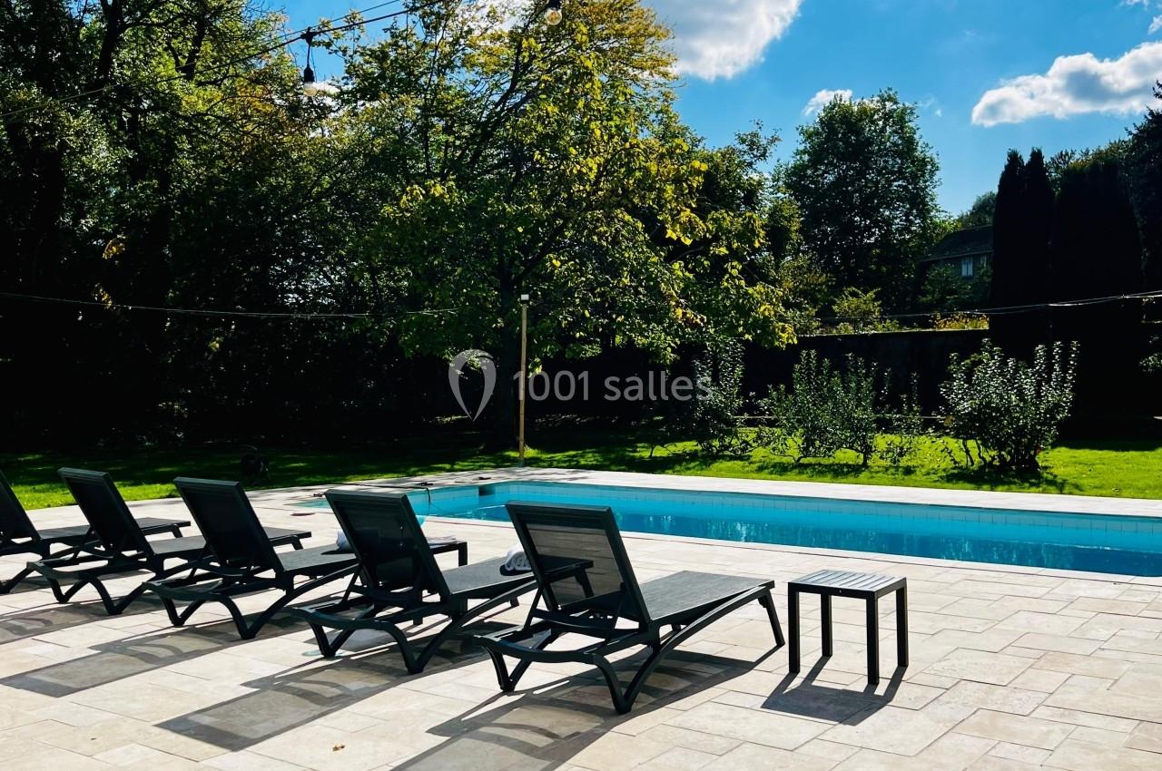 Chaises longues noires alignées près d'une piscine extérieure entourée de verdure sous un ciel ensoleillé.