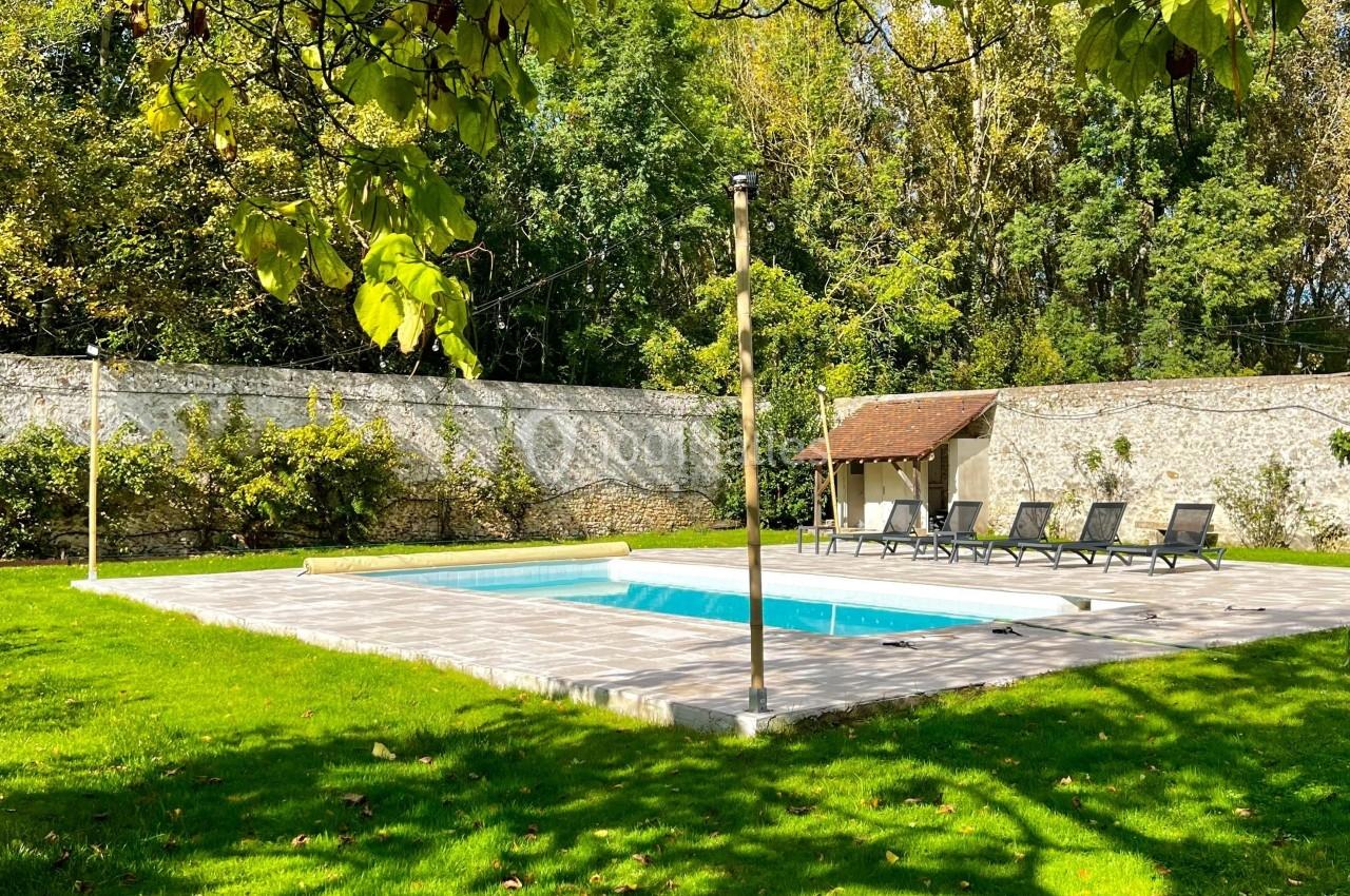 Piscine extérieure entourée de verdure, avec des transats alignés sur une terrasse en pierre et un mur en arrière-plan.