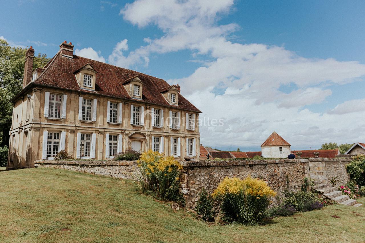 Grande maison en pierre avec toit en tuiles, entourée d'un jardin fleuri et d'un mur en pierre sous un ciel partiellement…