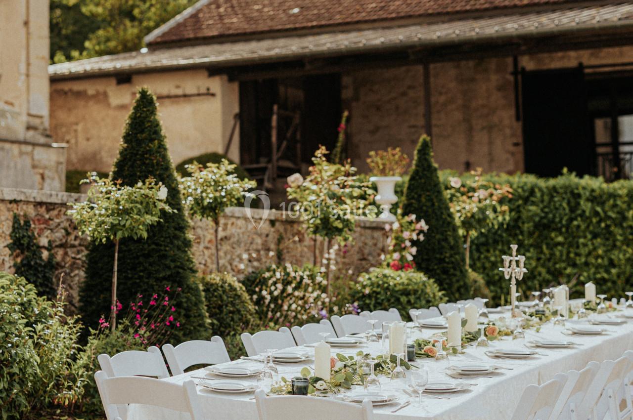 Table dressée en extérieur avec nappes blanches, chaises pliantes et décorations florales, devant un bâtiment ancien.