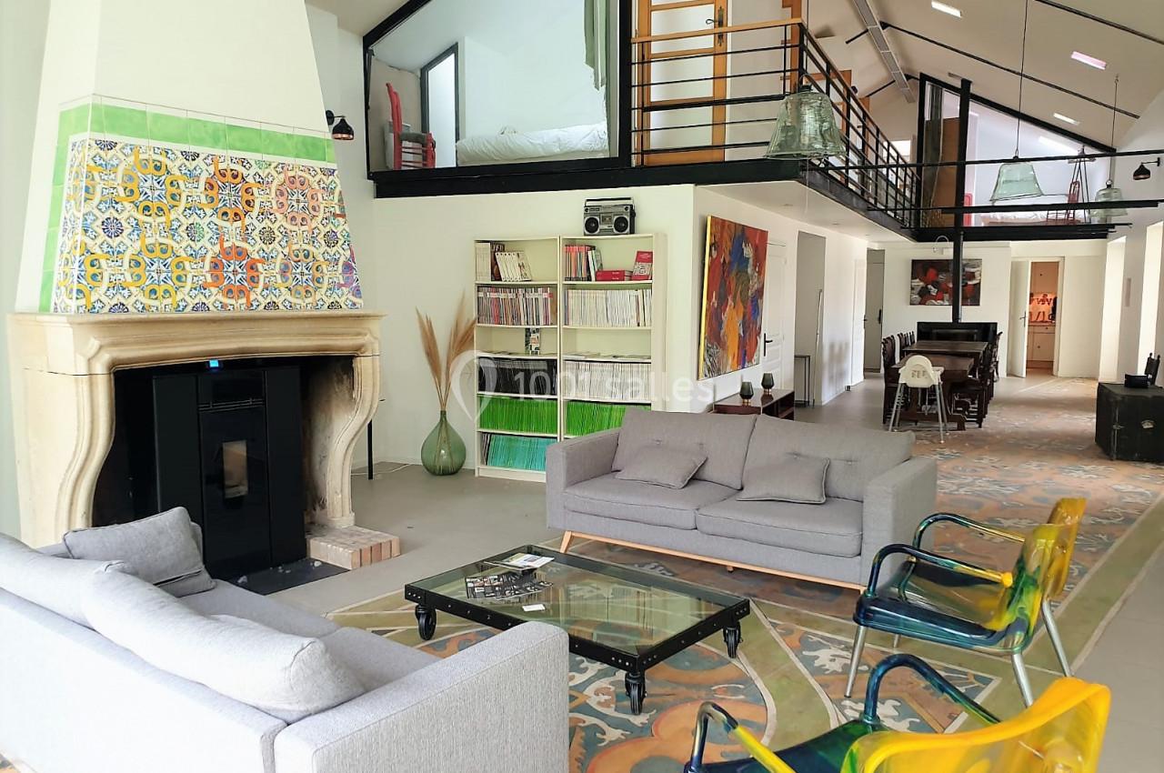 Salon spacieux avec cheminée ornée de carreaux colorés, canapés gris, chaises transparentes et mezzanine visible.