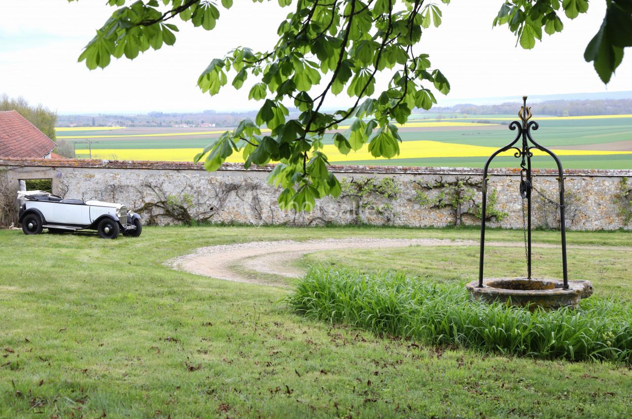 Voiture ancienne garée sur une pelouse près d'un puits en pierre, avec un mur en pierre et des champs en arrière-plan.