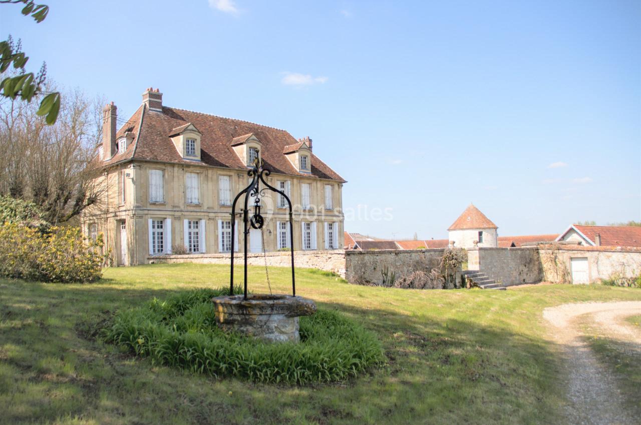 Grande maison en pierre avec toit en tuiles, puits ancien au premier plan et jardin verdoyant sous un ciel dégagé.