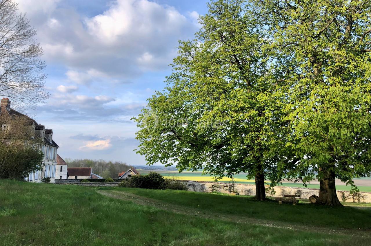 Maison ancienne près d'un grand arbre sur une pelouse verdoyante, avec un paysage champêtre en arrière-plan.