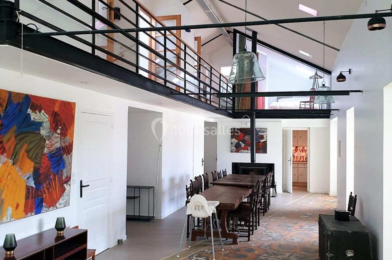 Intérieur lumineux avec mezzanine, grande table en bois, tapis coloré et œuvres d'art accrochées au mur.