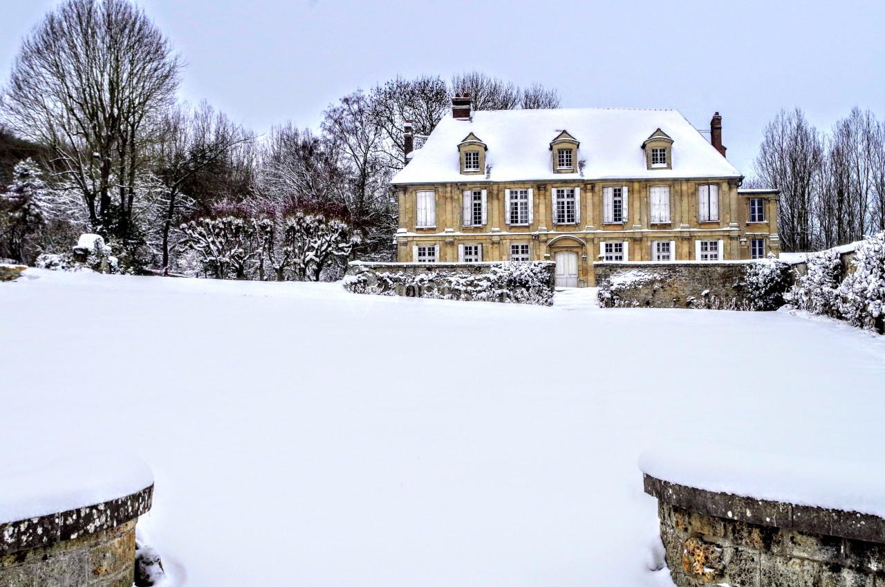 Manoir en pierre entouré d'un jardin recouvert de neige, avec des arbres dénudés en arrière-plan.