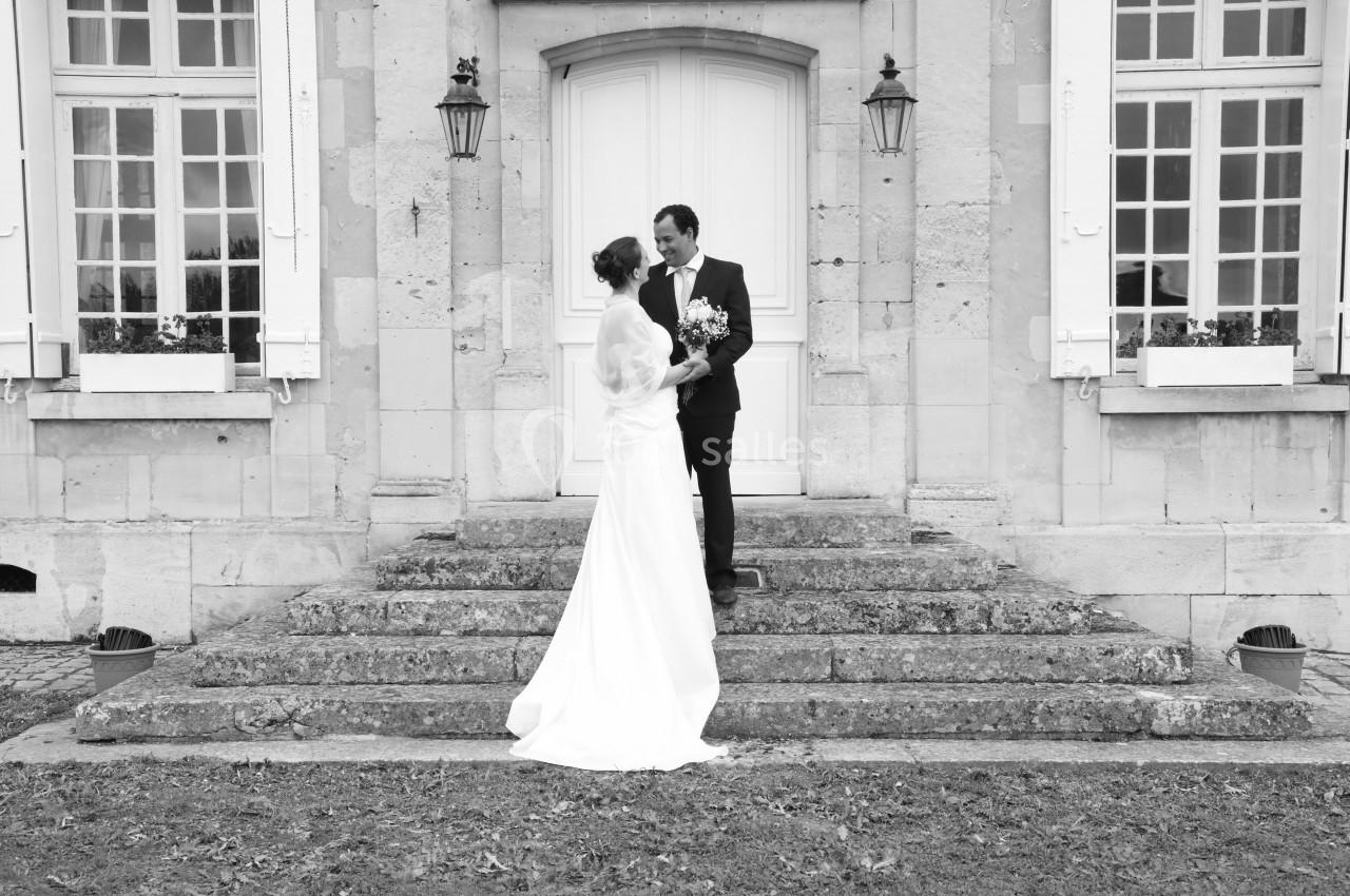 Un couple en tenue de mariage pose sur les marches devant une porte d'entrée d'un bâtiment en pierre.
