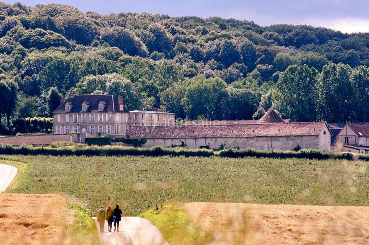 Un chemin bordé de champs mène à un grand bâtiment en pierre entouré de forêt sous un ciel partiellement nuageux.