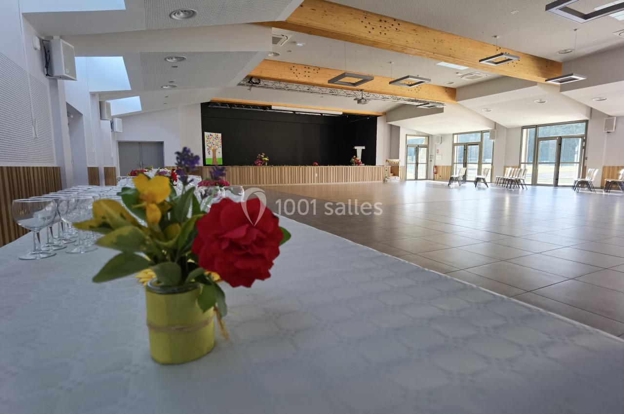 Salle lumineuse avec scène, tables décorées de fleurs au premier plan et grandes baies vitrées en arrière-plan.