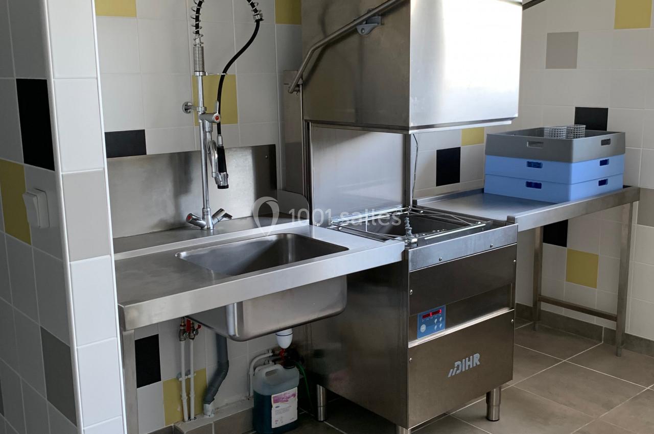 Évier en inox avec robinet professionnel, lave-vaisselle industriel et plans de travail dans une cuisine carrelée.