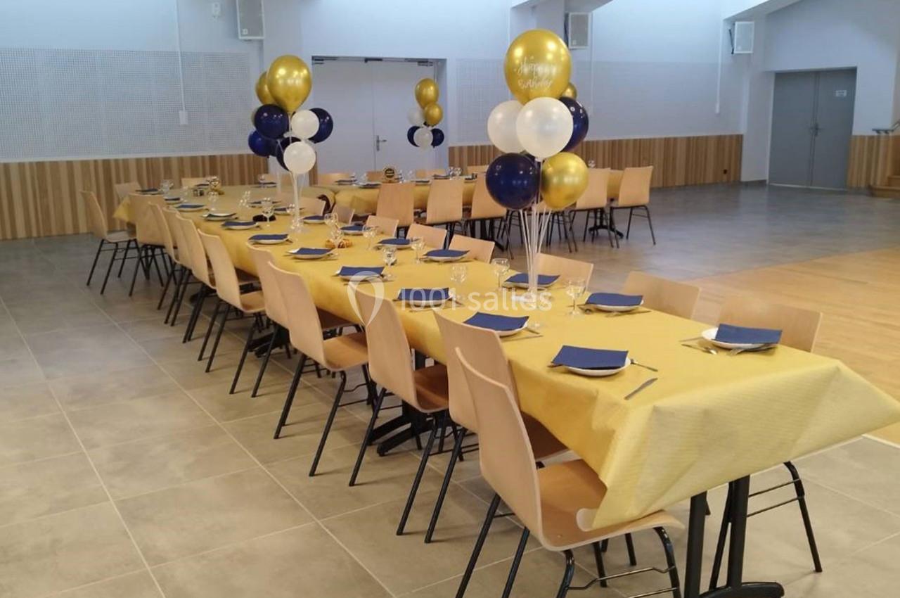 Tables alignées avec nappes jaunes, chaises beiges et décorations de ballons dorés, blancs et bleus dans une salle lumineuse.