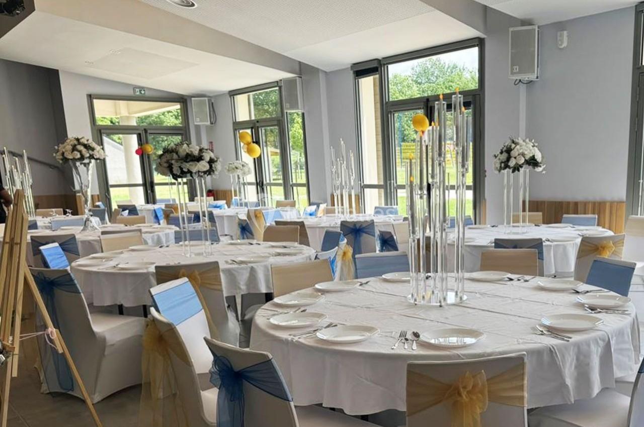 Salle de réception décorée avec des tables rondes, nappes blanches, chaises ornées de rubans colorés et centres de table…