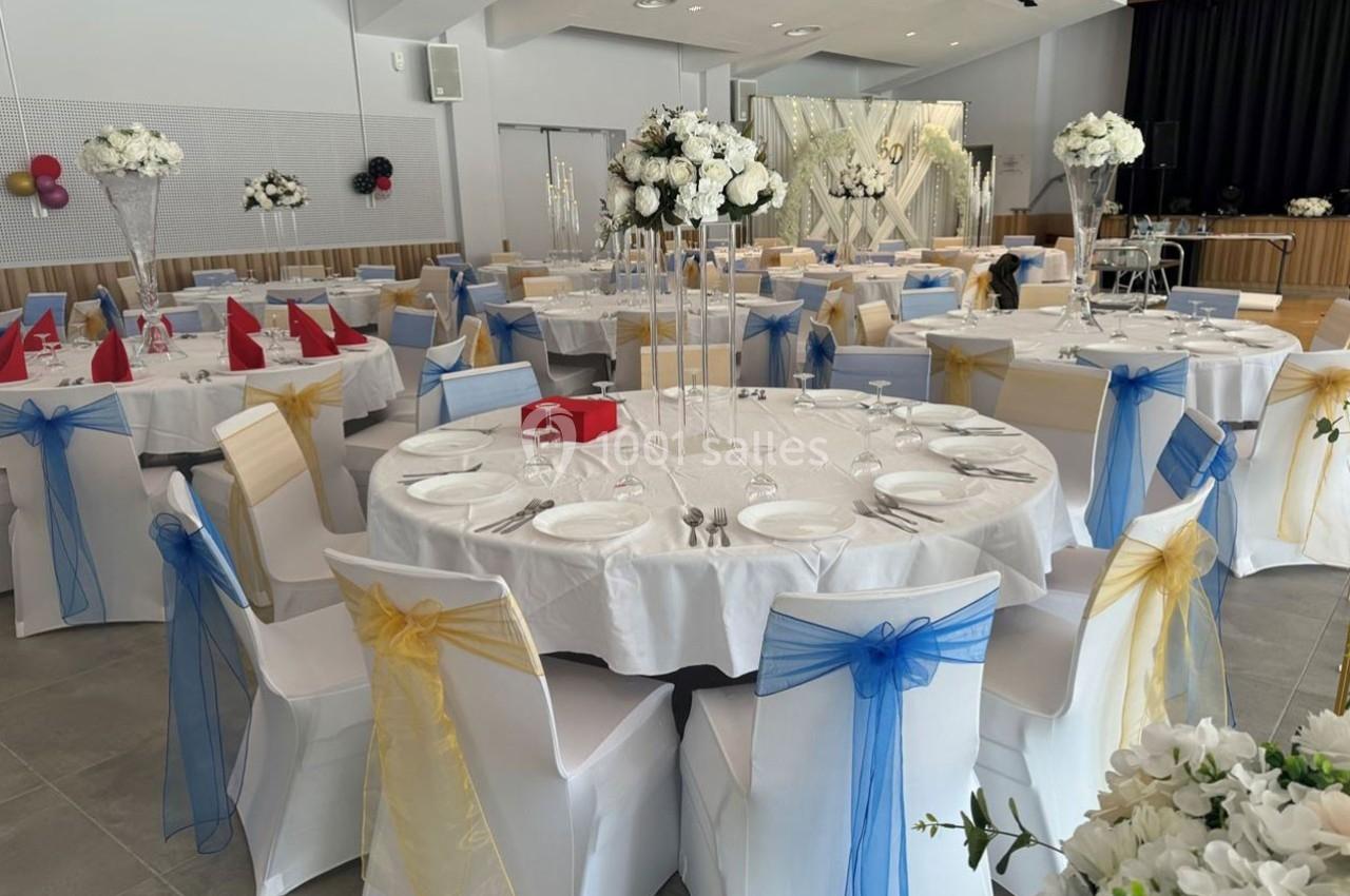 Salle de réception décorée avec des tables rondes, nappes blanches, chaises ornées de rubans colorés et centres de table…