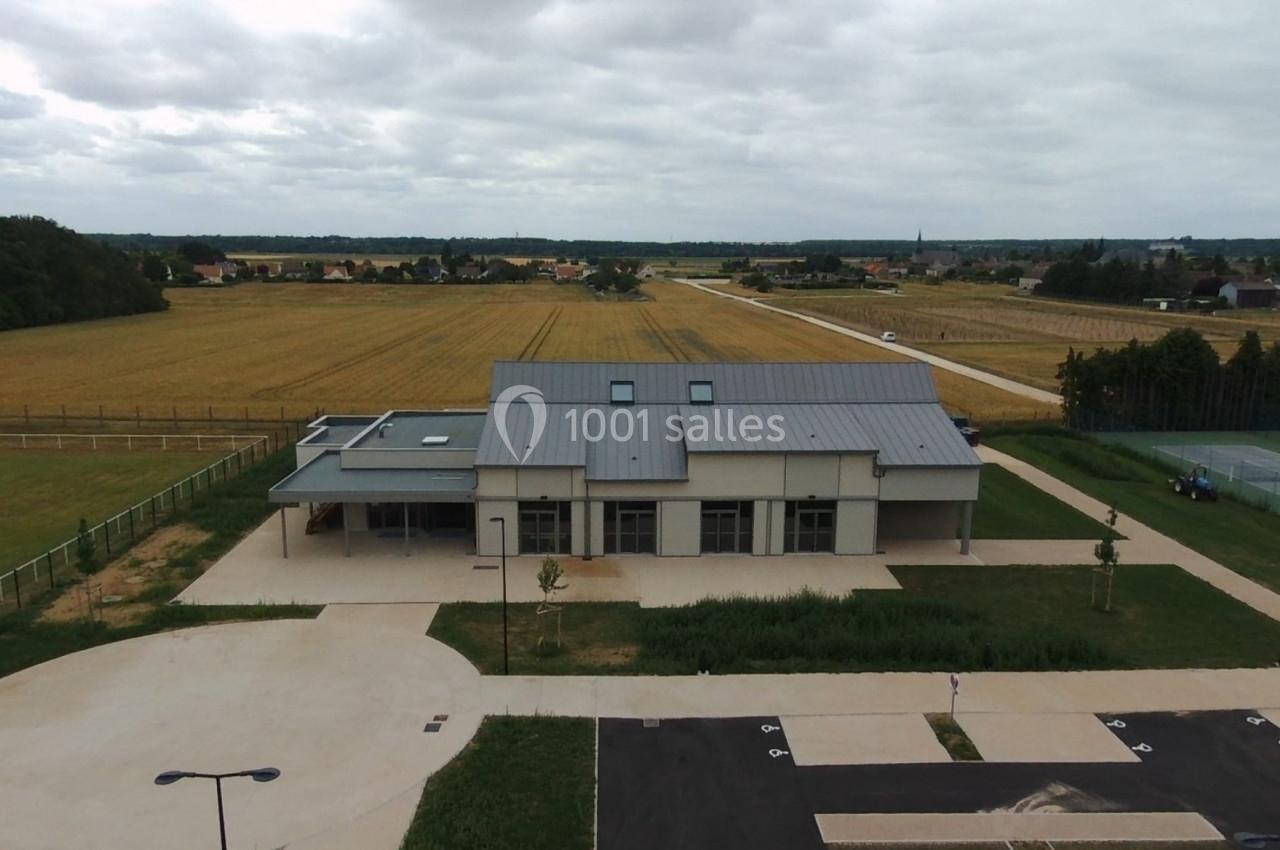 Vue aérienne d'un bâtiment moderne entouré de champs, avec un parking et des chemins aménagés à proximité.