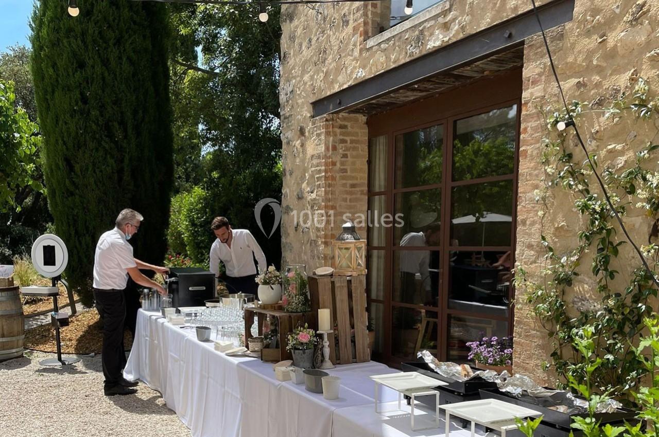 Deux serveurs préparent une table de buffet en extérieur, près d'un bâtiment en pierre entouré de végétation.