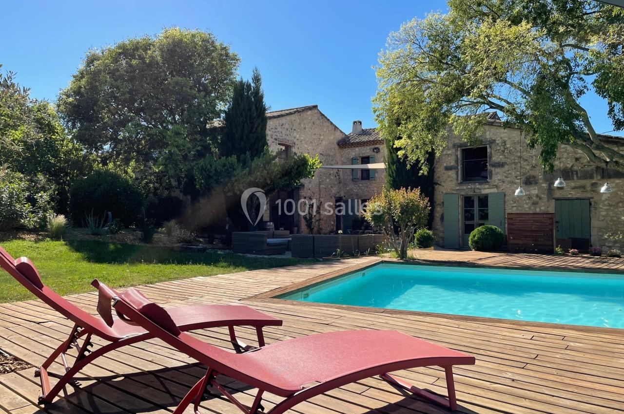 Deux chaises longues rouges sur une terrasse en bois près d'une piscine, avec une maison en pierre et des arbres en arrière…