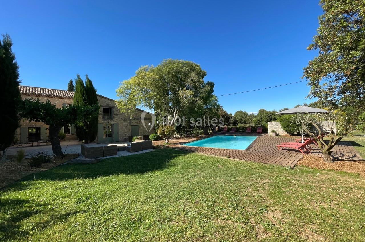 Jardin avec piscine, terrasse en bois, chaises longues rouges, parasol et maison en pierre sous un ciel bleu.
