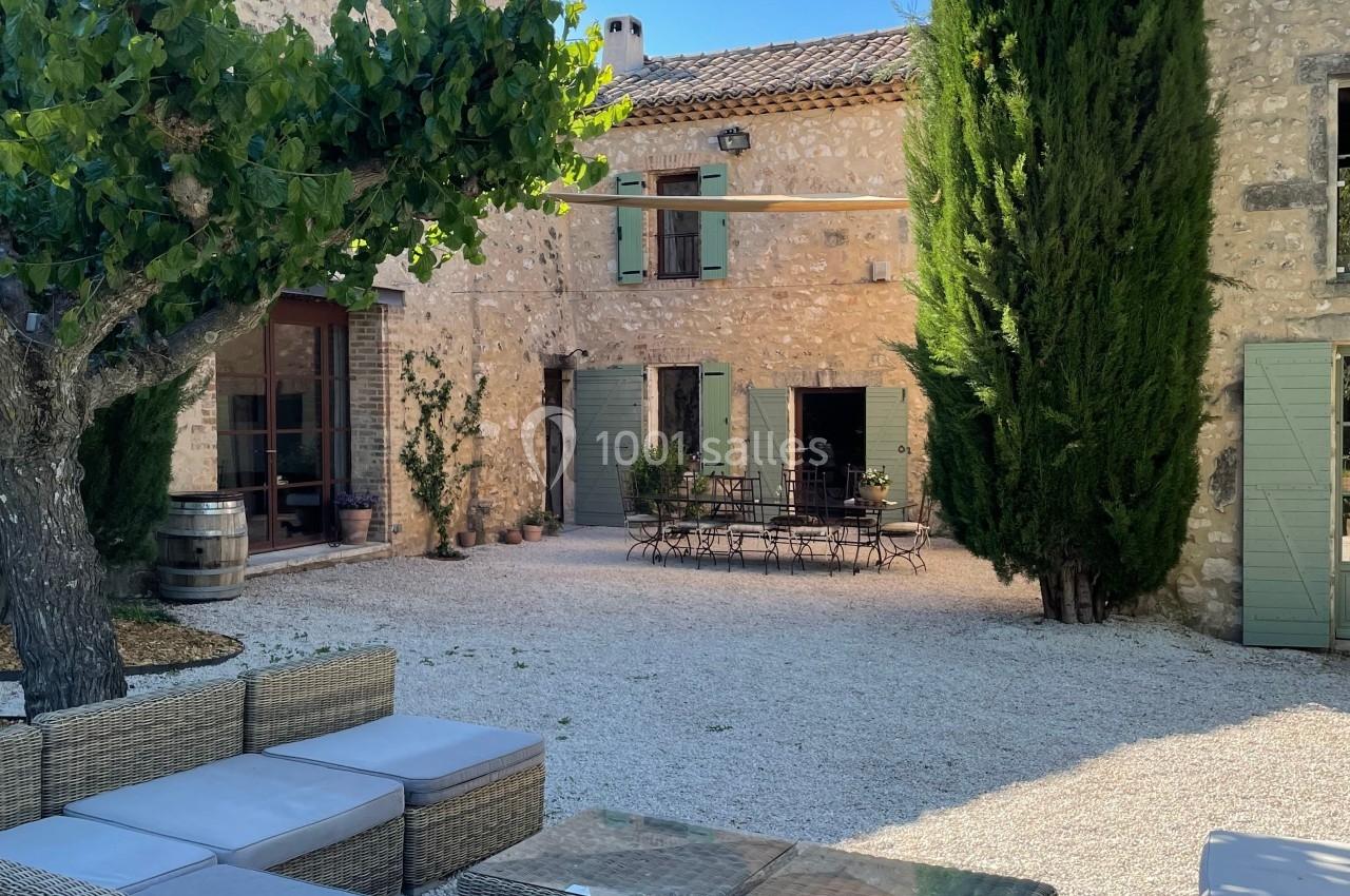 Cour en gravier avec mobilier en rotin, table et chaises en fer forgé, entourée de bâtiments en pierre et de cyprès.