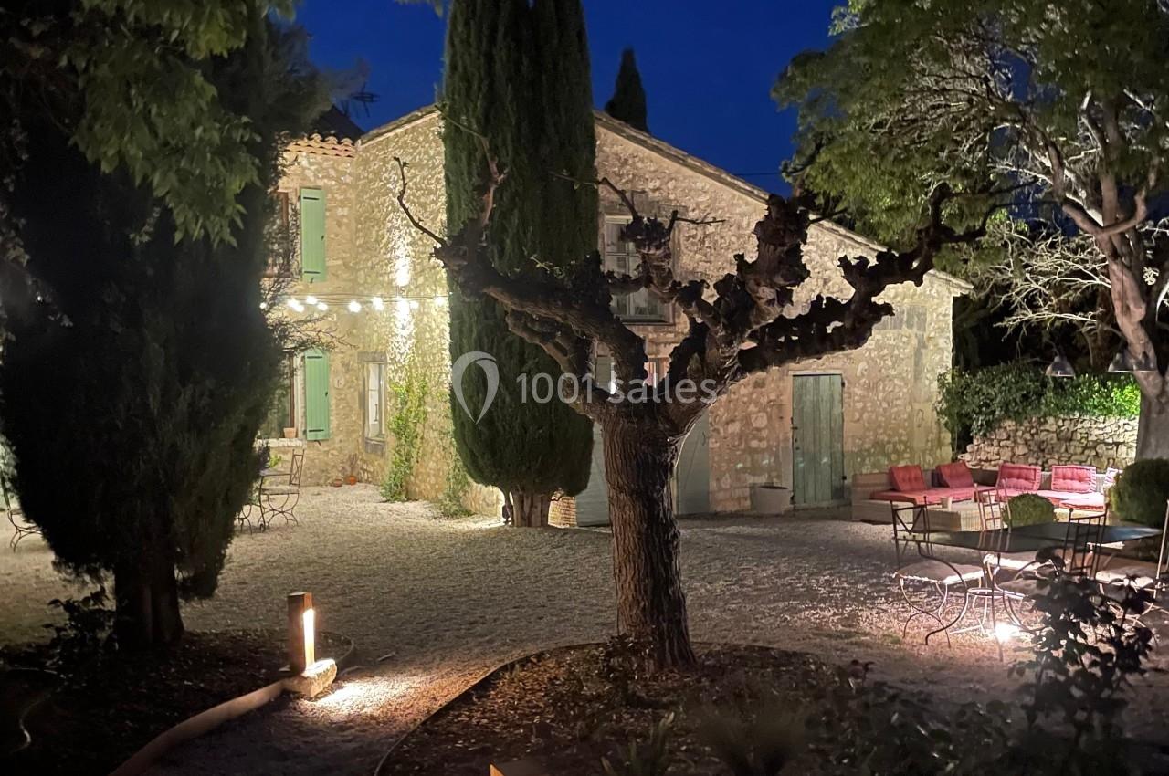 Cour d'une maison en pierre éclairée la nuit, avec arbres, mobilier de jardin et chemin bordé de lampes.