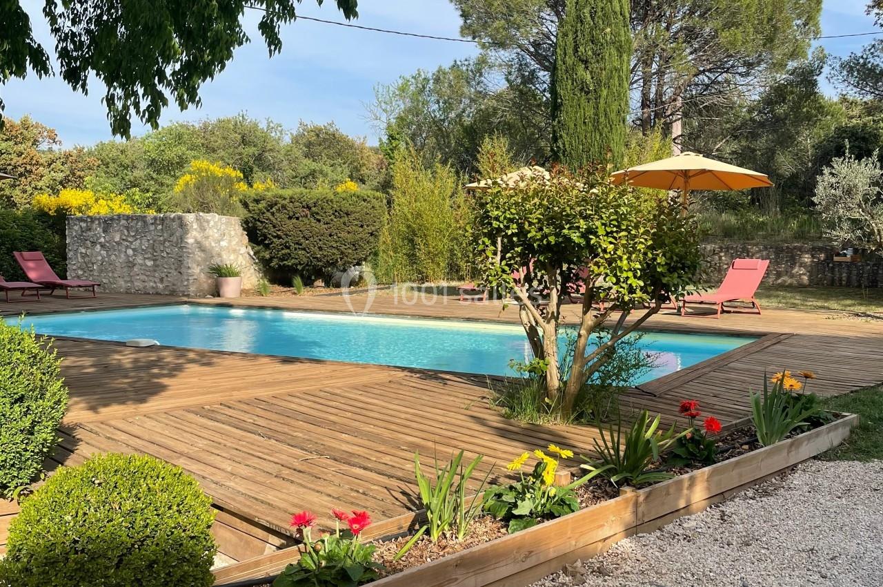Piscine extérieure entourée de végétation, terrasse en bois, transats rouges et parasol jaune sous un ciel dégagé.