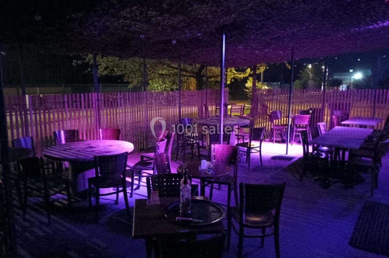 Terrasse extérieure d'un restaurant avec des tables et chaises en bois, éclairée par une lumière violette la nuit.