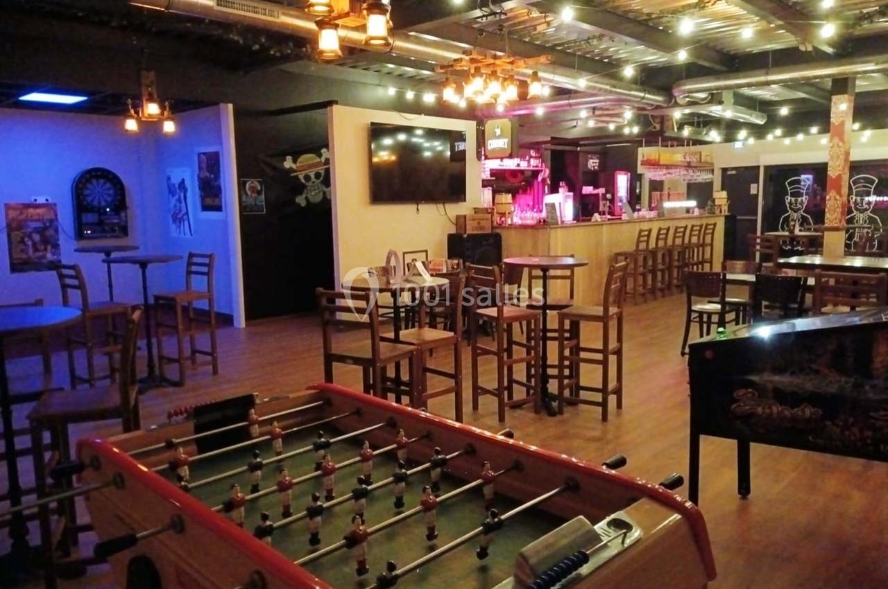 Salle de loisirs avec baby-foot, fléchettes, tables et bar éclairé dans une ambiance chaleureuse et industrielle.