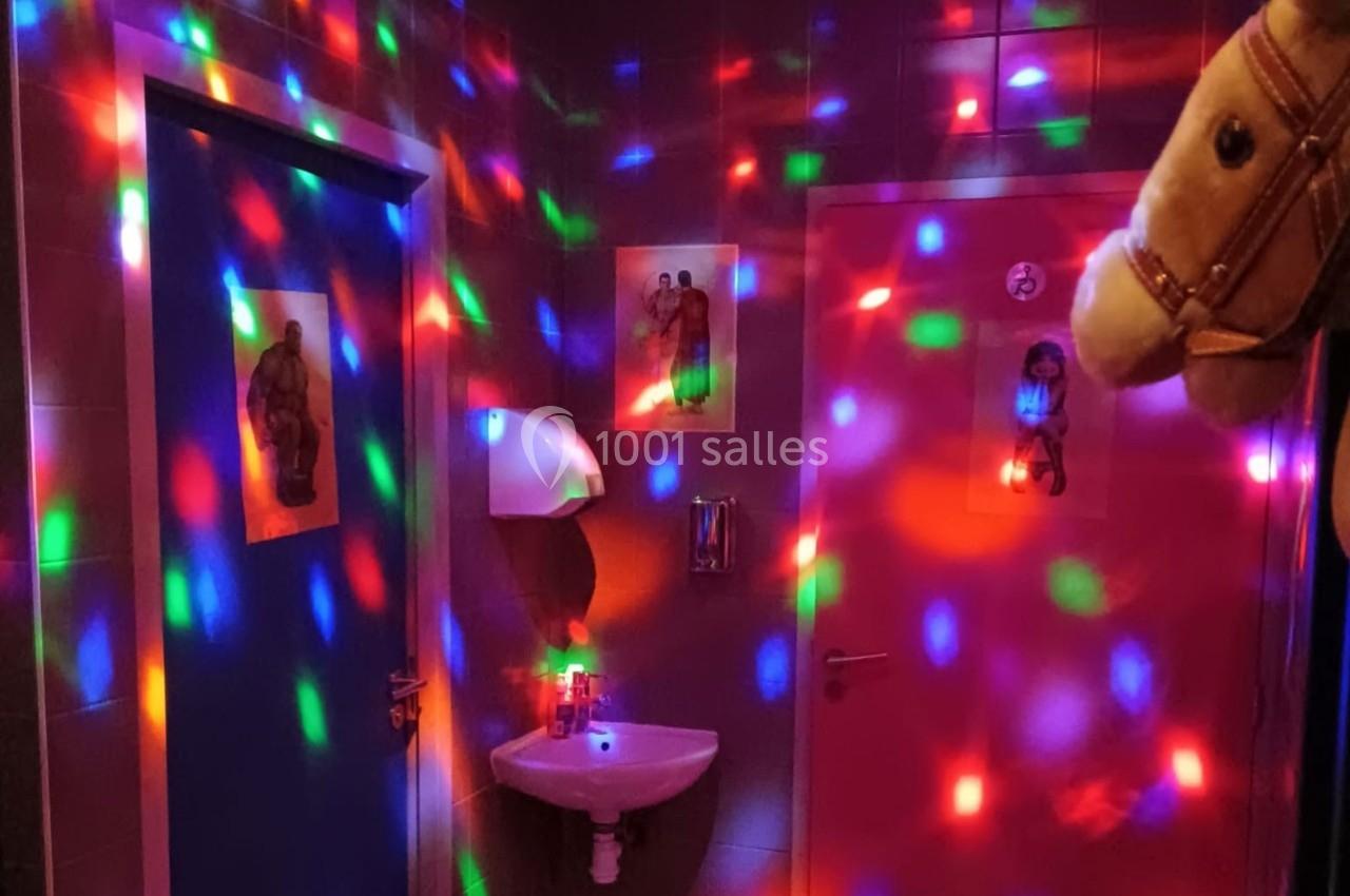 Évier mural dans une pièce sombre éclairée par des lumières colorées, avec deux portes et une tête de cheval en peluche.