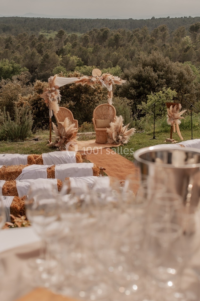 Chaises décorées sous une arche florale dans un cadre naturel, avec des rangées de bottes de paille en arrière-plan.