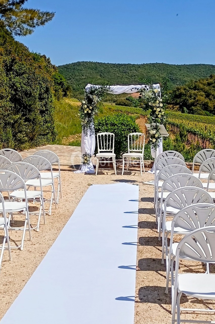 Allée centrale blanche menant à une arche de mariage décorée de fleurs, entourée de chaises blanches en extérieur.