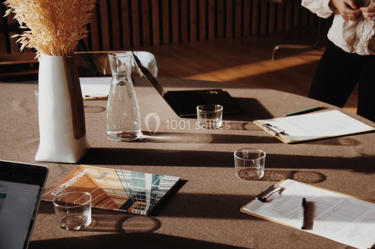 Table avec vase, carafe, verres, documents et carnets dans une salle lumineuse au mobilier en bois.