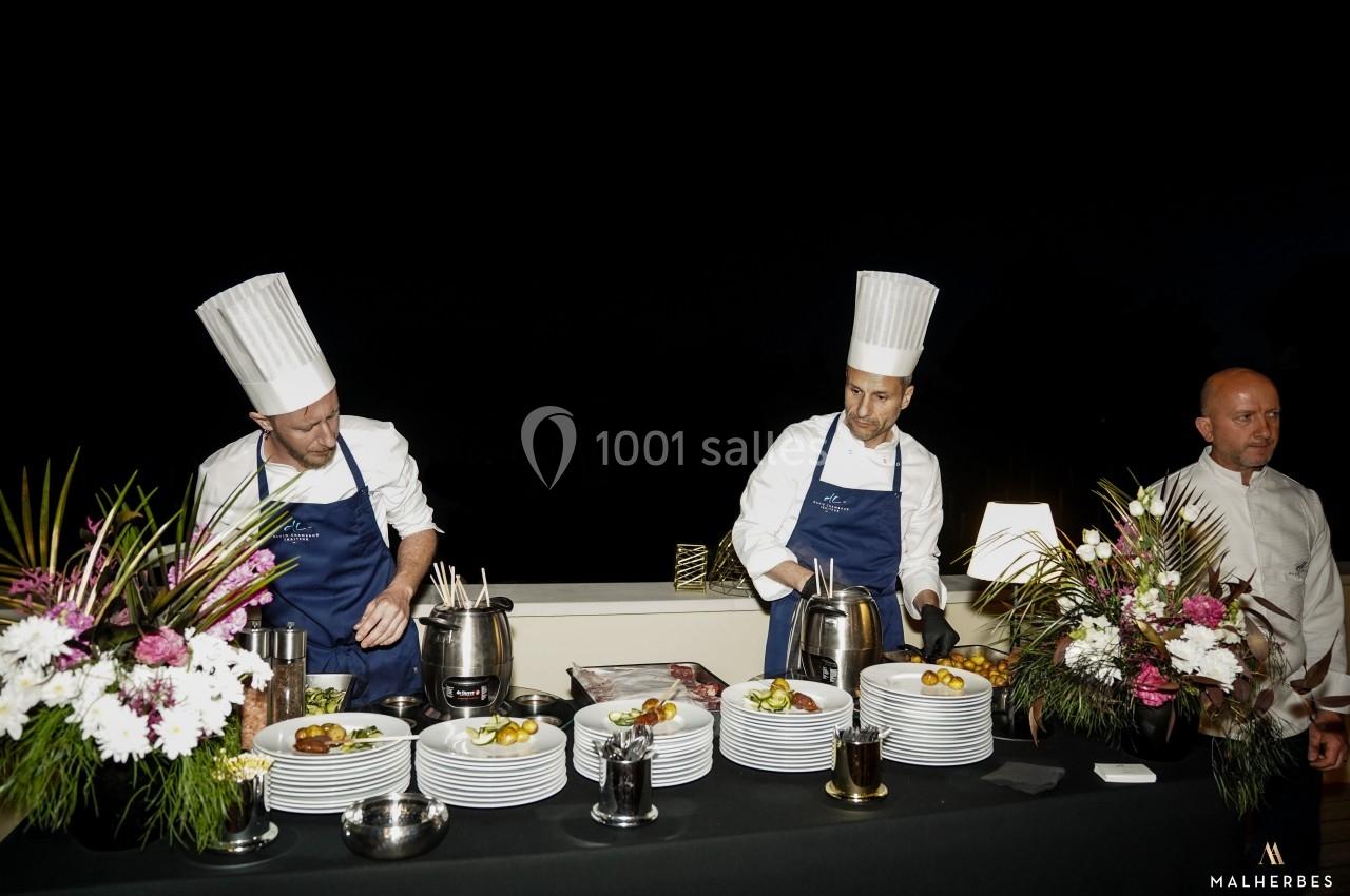 Deux chefs en tenue blanche et bleue préparent des plats sur une table décorée de fleurs, en extérieur de nuit.