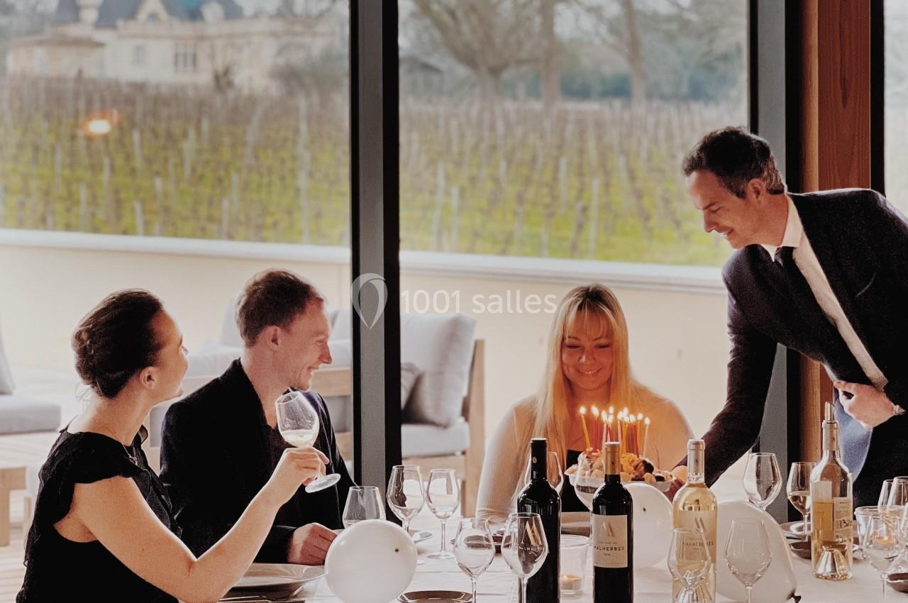 Un serveur apporte un dessert avec des bougies à une table où trois personnes dînent près d'une grande baie vitrée.