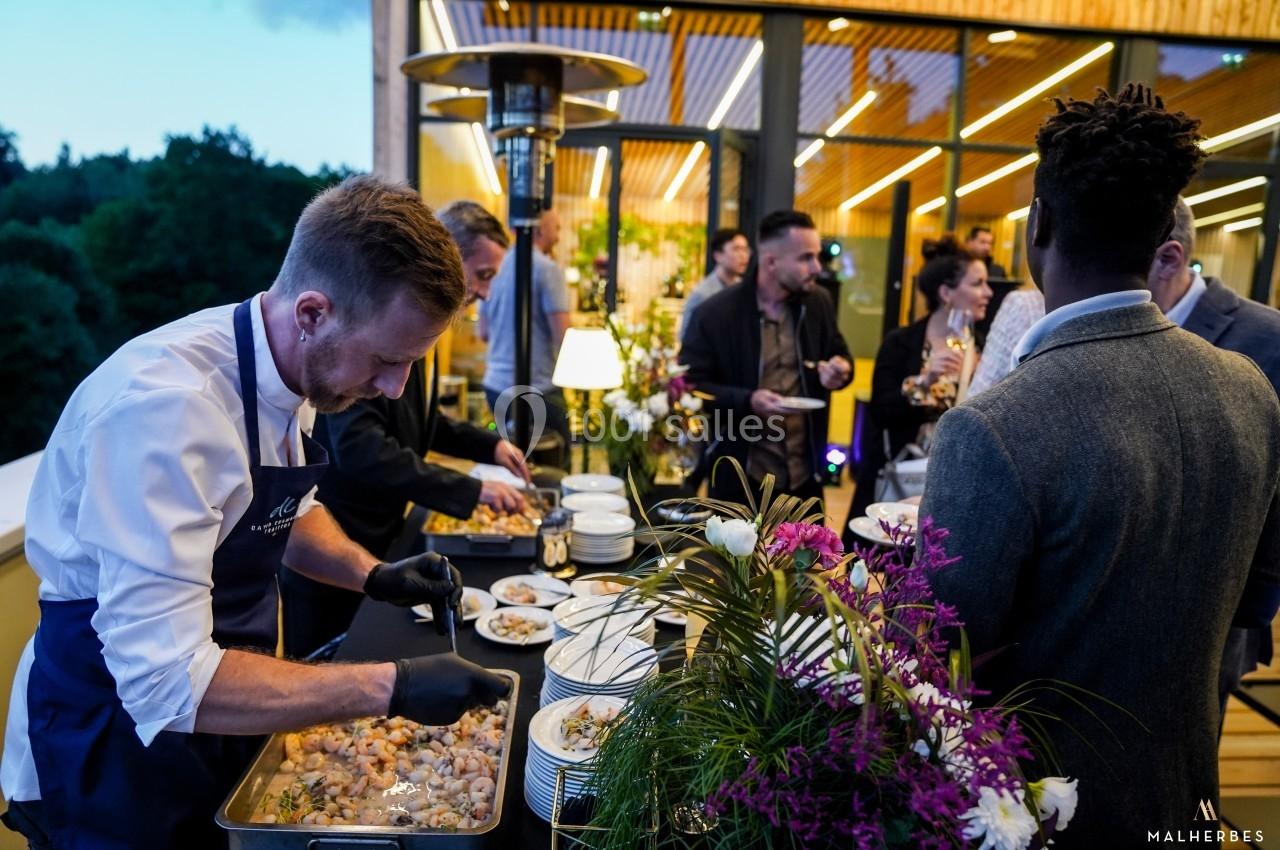 Un chef prépare des plats lors d'un événement en plein air, entouré d'invités et de décorations florales.