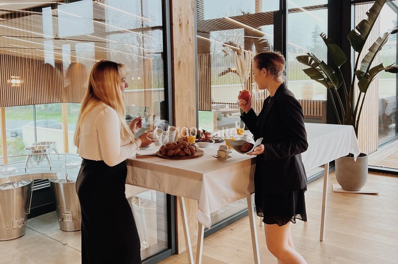 Deux femmes debout près d'une table dégustent un buffet dans un espace lumineux avec de grandes baies vitrées.