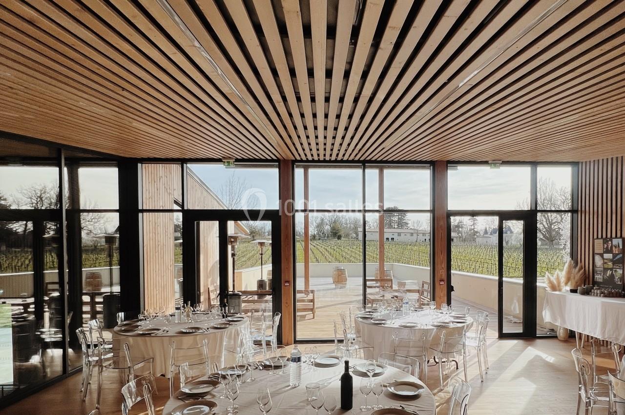 Salle lumineuse avec tables rondes dressées, chaises transparentes et grandes baies vitrées donnant sur un extérieur…