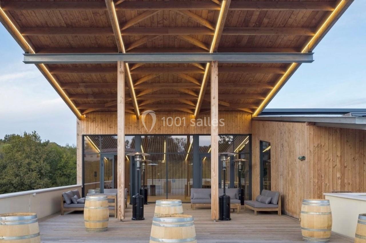 Terrasse en bois avec des tonneaux décoratifs, un toit moderne et des fauteuils devant une structure vitrée.