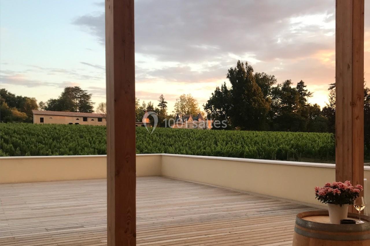 Terrasse en bois avec vue sur un vignoble au coucher du soleil, un tonneau et un pot de fleurs au premier plan.
