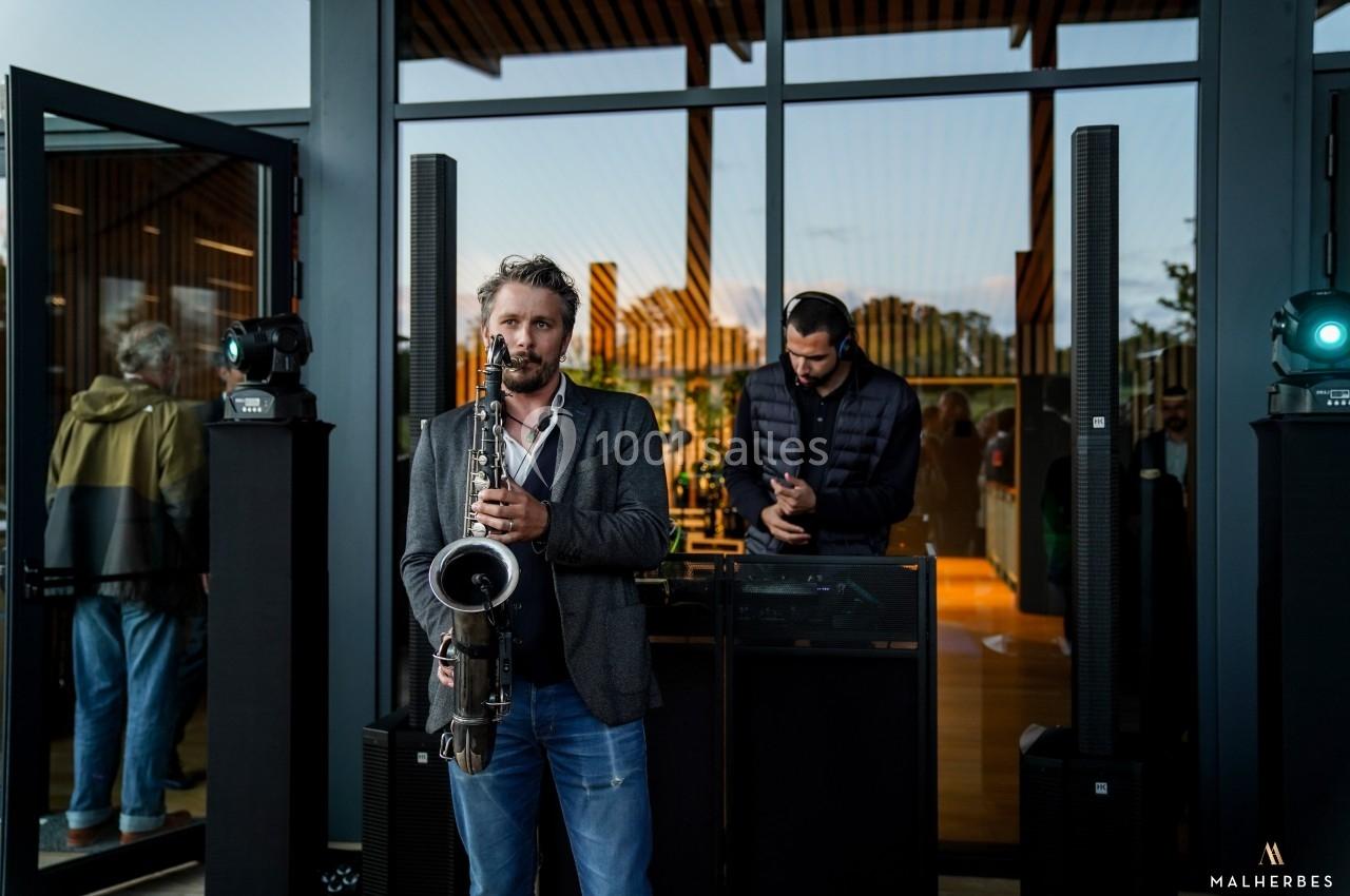 Un musicien joue du saxophone devant une baie vitrée, accompagné d'un DJ en arrière-plan.