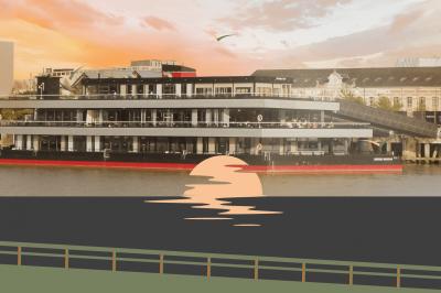 Terrasse avec tables, chaises et plantes, donnant sur une rivière bordée de bâtiments et d'un pont au loin.