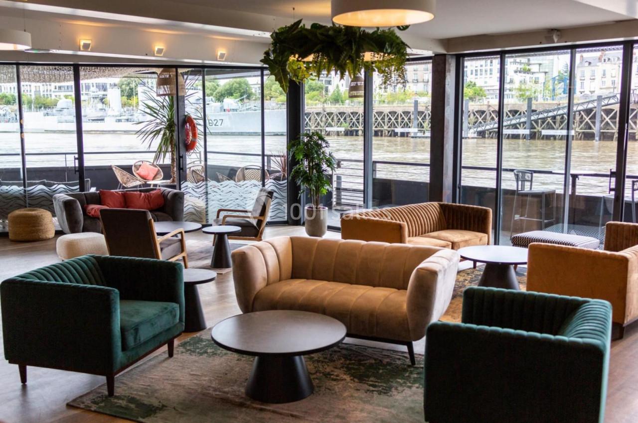 Espace lounge avec fauteuils colorés, tables basses et grandes baies vitrées offrant une vue sur un fleuve et un quai.