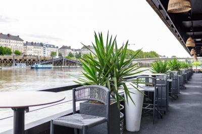 Terrasse avec tables, chaises et plantes, donnant sur une rivière bordée de bâtiments et d'un pont au loin.