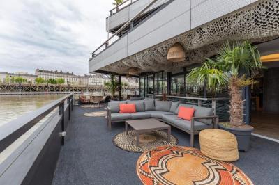 Terrasse avec tables, chaises et plantes, donnant sur une rivière bordée de bâtiments et d'un pont au loin.