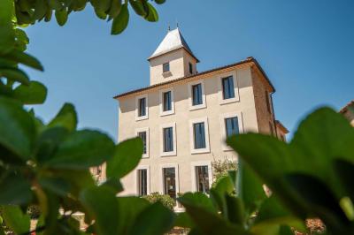 Façade d'un bâtiment clair avec une terrasse aménagée, un grand arbre et un ciel bleu en arrière-plan.