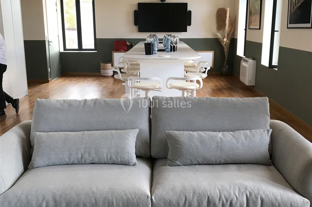 Canapé gris clair devant une grande table blanche entourée de chaises, dans une pièce lumineuse au sol en bois.