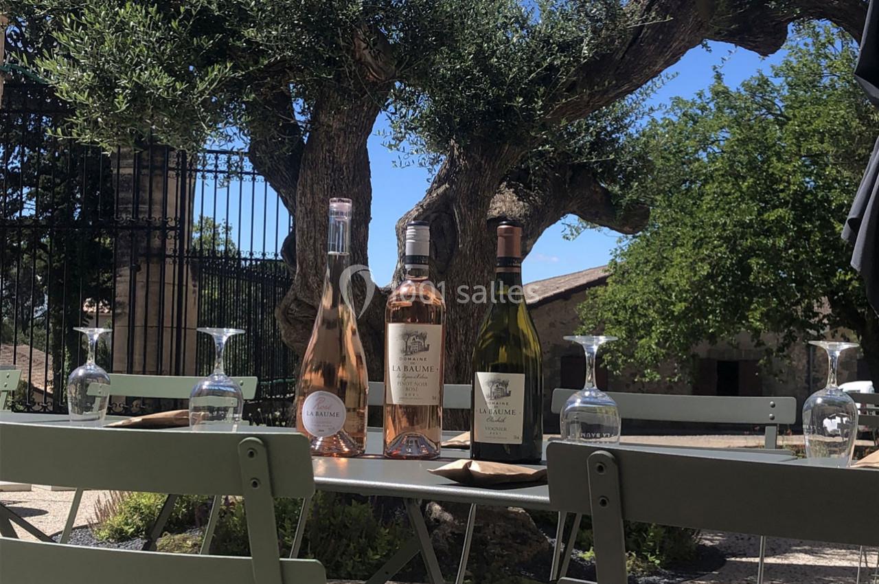 Bouteilles de vin et verres disposés sur une table en extérieur, sous des oliviers, par une journée ensoleillée.