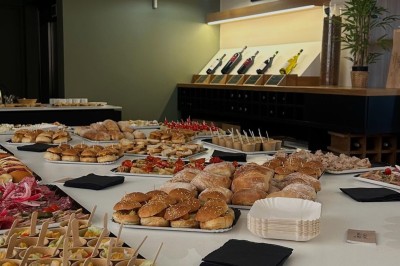 Salle lumineuse avec un long comptoir blanc, des tabourets, des bouteilles exposées et des poutres apparentes au plafond.