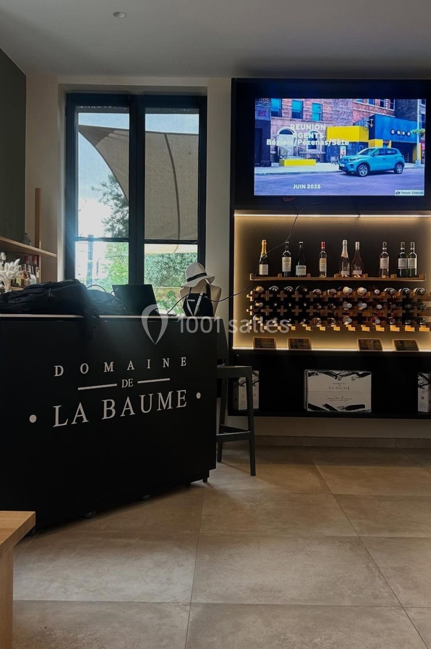Comptoir noir avec inscription ’Domaine de La Baume’, étagères de bouteilles de vin et écran affichant une publicité.