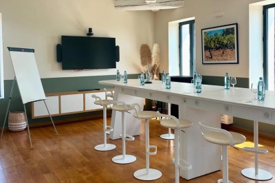 Salle lumineuse avec un long comptoir blanc, des tabourets, des bouteilles exposées et des poutres apparentes au plafond.