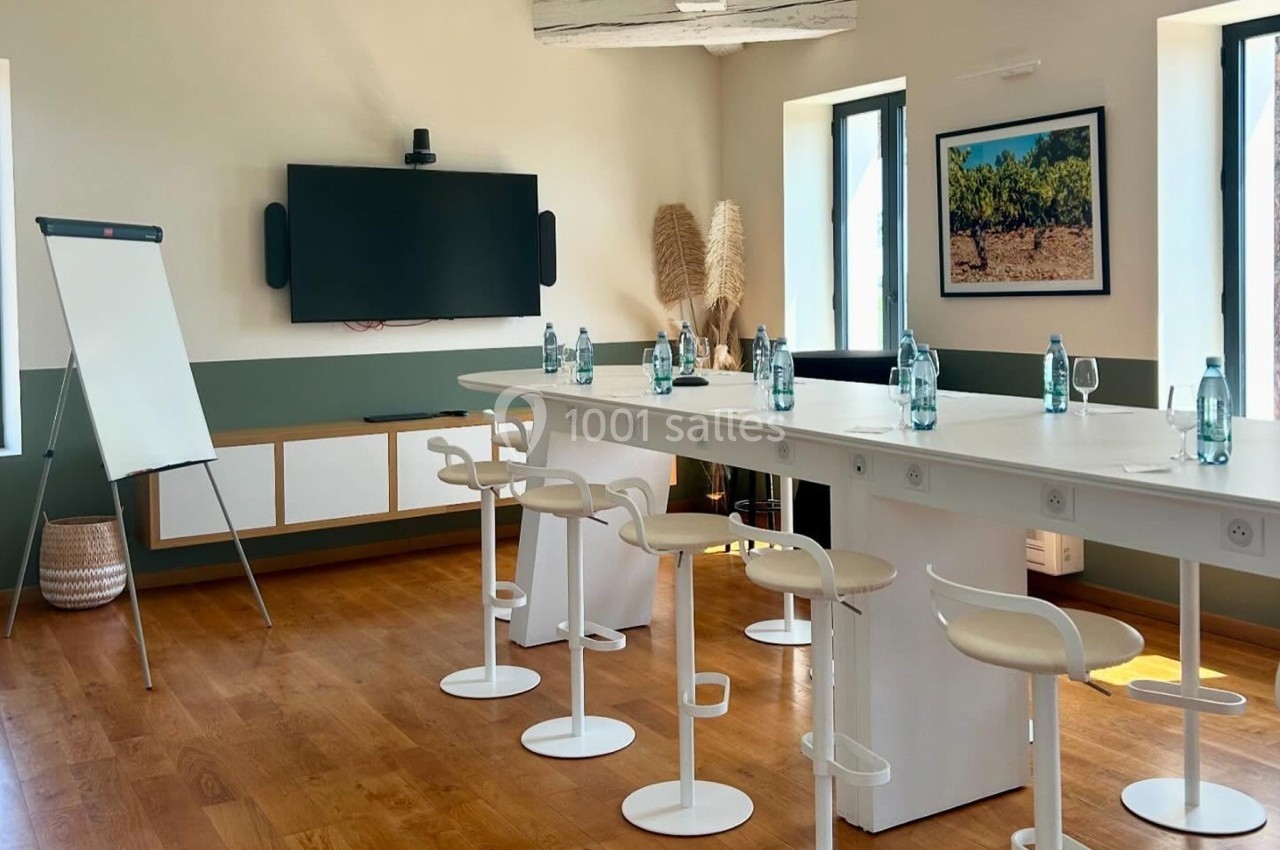 Salle de réunion lumineuse avec table blanche, chaises hautes, écran mural, tableau blanc et bouteilles d'eau.