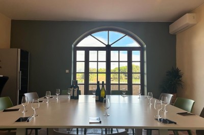 Salle lumineuse avec un long comptoir blanc, des tabourets, des bouteilles exposées et des poutres apparentes au plafond.